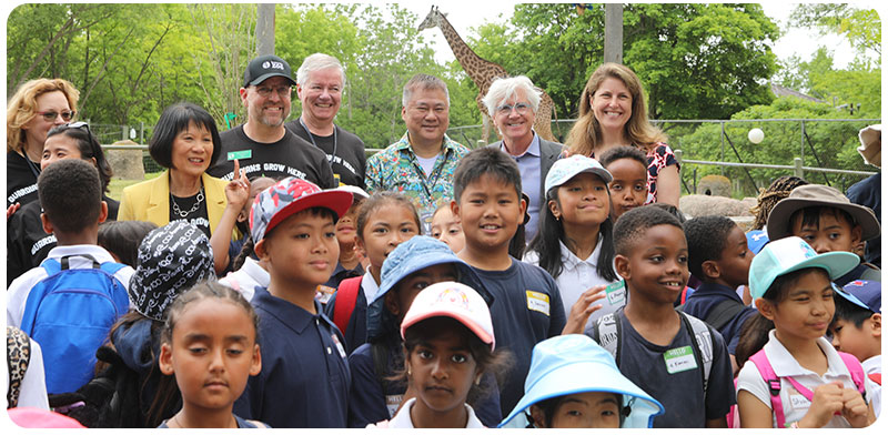 Group shot of speakers and school kids with giraffes walking in the background.